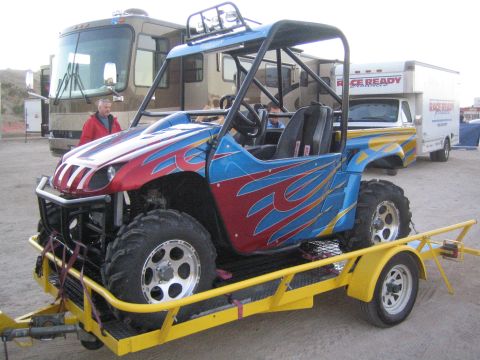 1990 Ford Bronco - red, white and blue UTV side-by-side with flame graphics strapped to a yellow utility trailer in a desert lot.