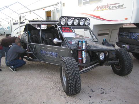 1990 Ford Bronco - black tube-chassis off-road buggy with roll cage and For Sale sign, parked beside a fifth-wheel RV in the dirt.