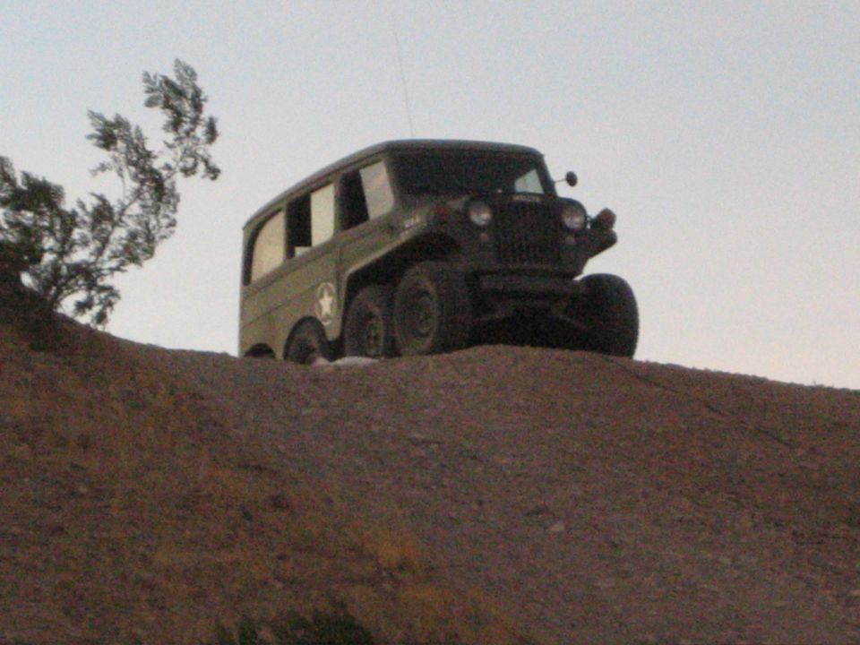 Olive-green military-style six-wheeled jeep parked atop a dirt hill at dusk
