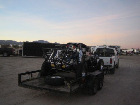 1990 Ford Bronco - white pickup truck towing a tandem-axle trailer carrying an off-road tube-chassis buggy in a dusty lot at dusk.