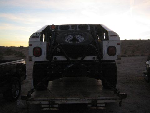 1990 Ford Bronco - Rear view of a white tube-bodied off-road race vehicle with spare tire mounted, loaded on a flatbed trailer at dusk.