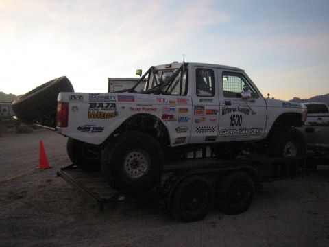 1990 Ford Bronco - white off-road prerunner race truck covered in sponsor decals and numbered 1500, loaded on a trailer in the desert at dusk.