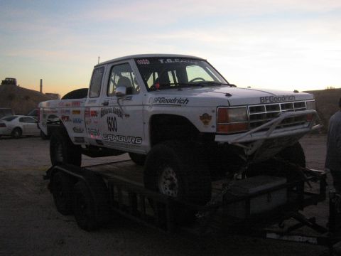 1990 Ford Bronco - white lifted off-road race truck numbered 1500 with BFGoodrich and Camburg sponsor decals, parked on a trailer at dusk.