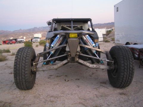 1990 Ford Bronco - Front view of a tube-chassis off-road race buggy with exposed A-arm suspension and blue bypass shocks, parked on dirt at dusk.