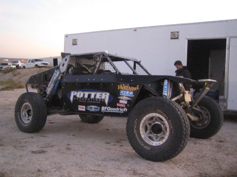 1990 Ford Bronco - black tube-chassis off-road buggy with Potter Productions and BFGoodrich decals parked beside a white enclosed trailer.
