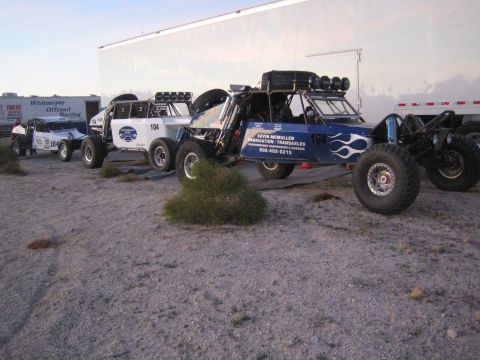 1990 Ford Bronco - Several blue and white off-road race buggies lined up beside enclosed trailers in a desert pit area at dusk.