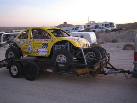 1990 Ford Bronco - yellow VW Beetle-bodied Baja Bug off-road racer numbered 500 with sponsor decals, loaded on a tandem-axle trailer at dusk.