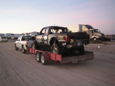 1990 Ford Bronco - white pickup towing a red trailer carrying a black off-road race truck with sponsor decals, on a dirt lot at dusk.