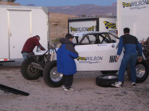 1990 Ford Bronco - Three people tend to a white off-road buggy marked Wilson Motorsports 1614 beside enclosed trailers in a desert lot.