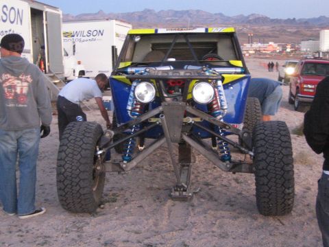 1990 Ford Bronco - Crew members work on a blue and yellow tube-chassis off-road buggy with exposed suspension, near Wilson Motorsports trailers at dusk.