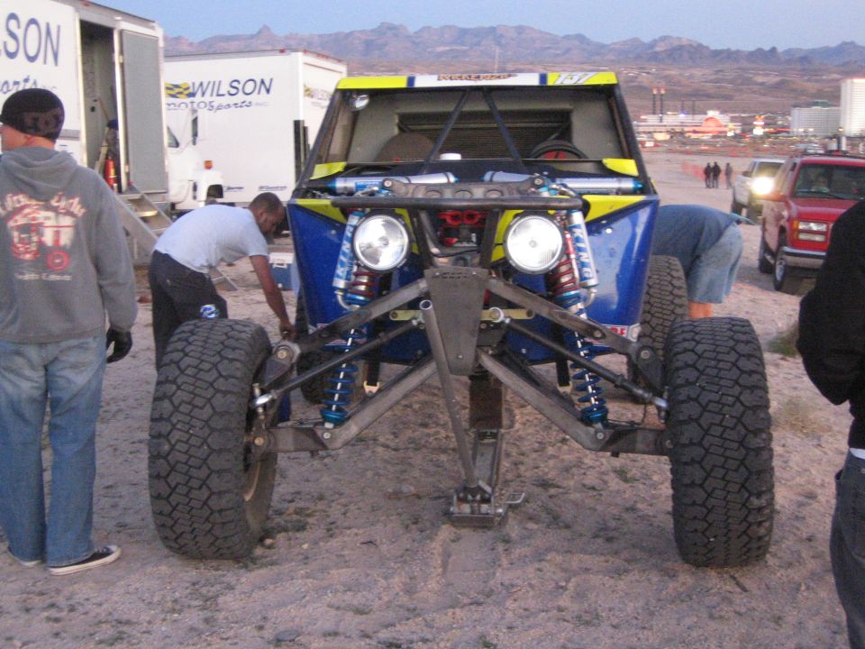 Front of a blue and yellow off-road buggy with crew working on tires in the desert