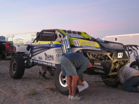 1990 Ford Bronco - man bends to inspect a blue and yellow off-road buggy numbered 137 with sponsor decals in a desert pit area at dusk.