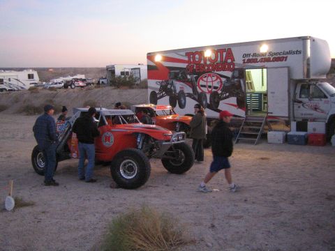 1990 Ford Bronco - At dusk, two orange off-road race trucks sit beside a Toyota of Escondido Off-Road Specialists box trailer as crew and spectators gather in…