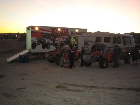1990 Ford Bronco - Off-road race buggies and crew gather beside a Toyota Off-Road Specialists box trailer and a motorhome in a dim desert pit at dusk.