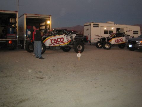 1990 Ford Bronco - Two TSCO Tube Specialties Company off-road race trucks sit beside an open box trailer and an RV in a dirt pit at dusk.