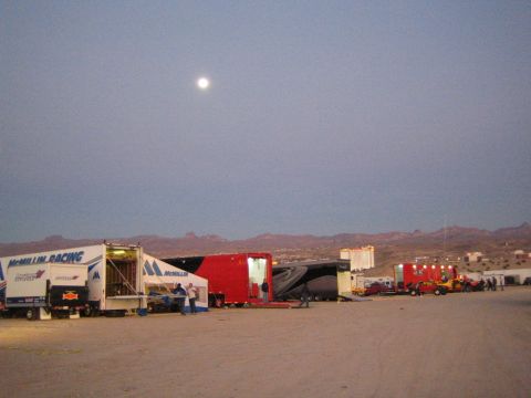 1990 Ford Bronco - row of race-team haulers, including a McMillin Racing trailer, lines a desert race camp under a rising full moon at dusk.