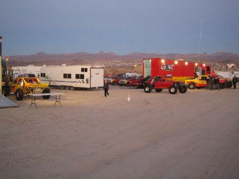 1990 Ford Bronco - Yellow off-road race trucks and crew gather beside haulers in a desert race pit at dusk, with a town and rugged hills in the background.