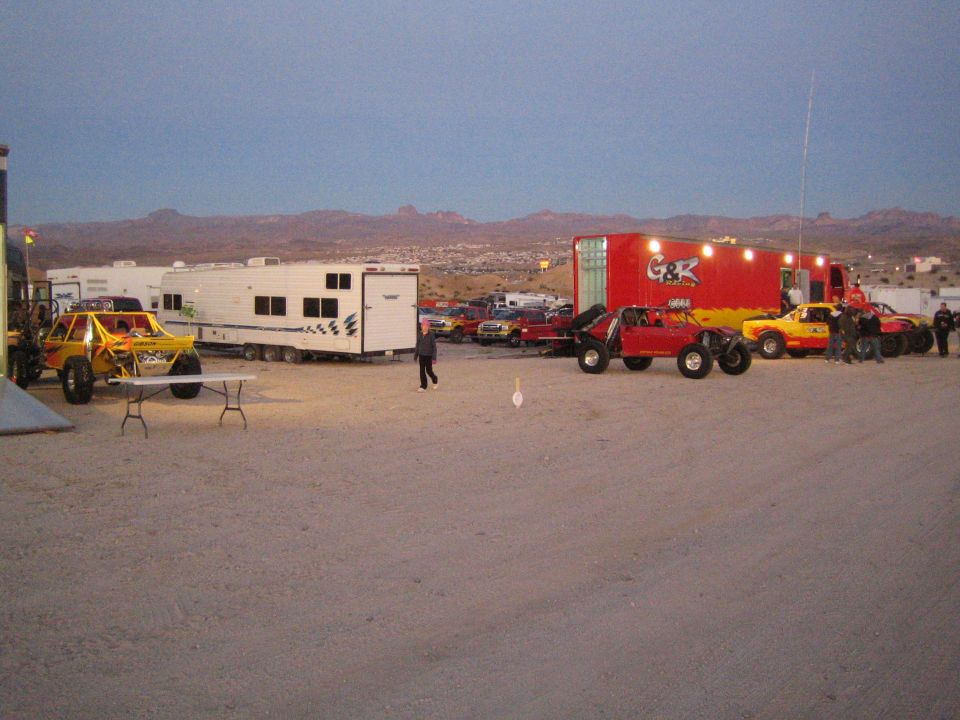 Yellow off-road race trucks beside trailers in a desert pit at dusk with town and hills behind