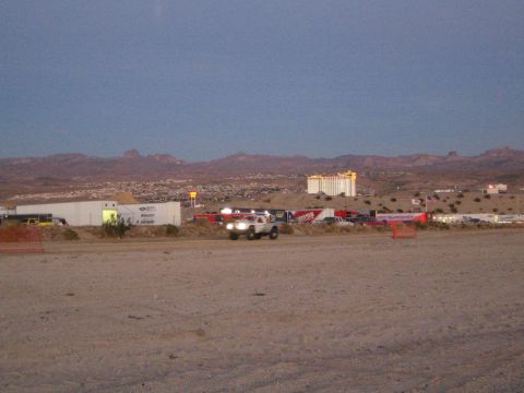 1990 Ford Bronco - off-road truck with headlights blazing crosses a dirt course at dusk, with a tall casino tower and desert hills in the distance.