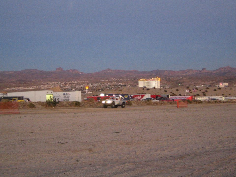 Off-road truck with headlights on a dirt course at dusk near a casino tower and desert hills