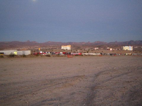 1990 Ford Bronco - wide dirt lot at dusk holds distant rows of race trailers and vehicles against a backdrop of buildings and desert hills.