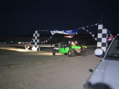 1990 Ford Bronco - bright green sand rail and another buggy line up under a checkered-flag start banner strung with pennants at night.