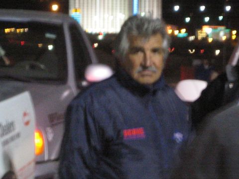 1990 Ford Bronco - gray-haired, mustached man in a blue jacket stands beside a white truck at night with blurred city lights behind him.