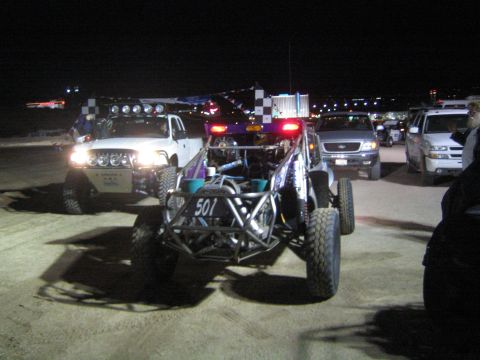 1990 Ford Bronco - purple tube-frame off-road buggy marked 501 sits among pickup trucks with headlights on in a dirt staging area at night.