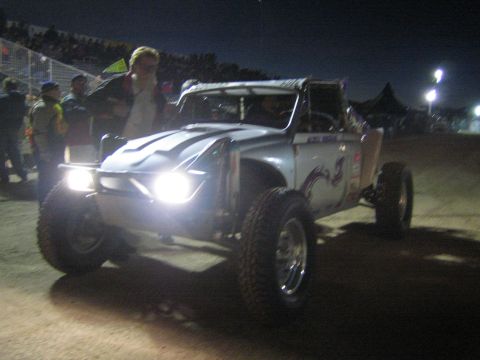 1990 Ford Bronco - white off-road race truck with its headlights blazing rolls past a crowd of spectators in a dirt arena at night.