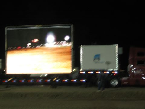 1990 Ford Bronco - large mobile LED video screen mounted on a trailer displays race footage beside a semi at a night dirt-racing event.