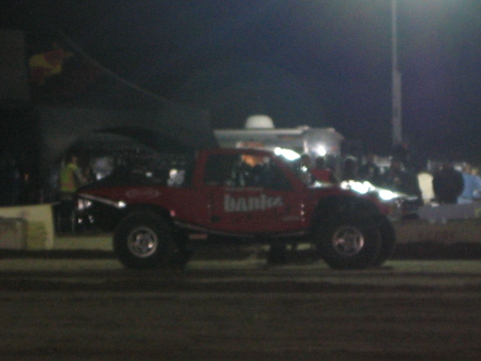 Red Banks off-road truck on a dirt course at night with stadium crowd behind