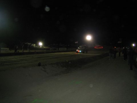 1990 Ford Bronco - dark dirt race course at night shows a distant vehicle and scattered bright lights, with spectators along the far edge.