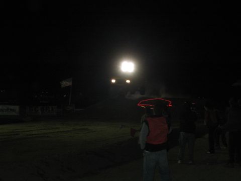 1990 Ford Bronco - spectator in a red vest watches a vehicle with glaring lights approach across a dark dirt course at night near SCORE banners.
