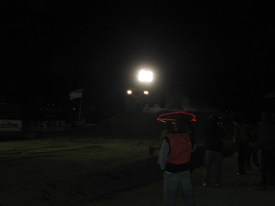 Spectator in red vest watching a glaring-light vehicle approach on a dirt course at night