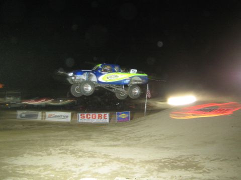 1990 Ford Bronco - blue-and-yellow off-road race truck launches off a dirt jump at night in front of SCORE and BFGoodrich banners.
