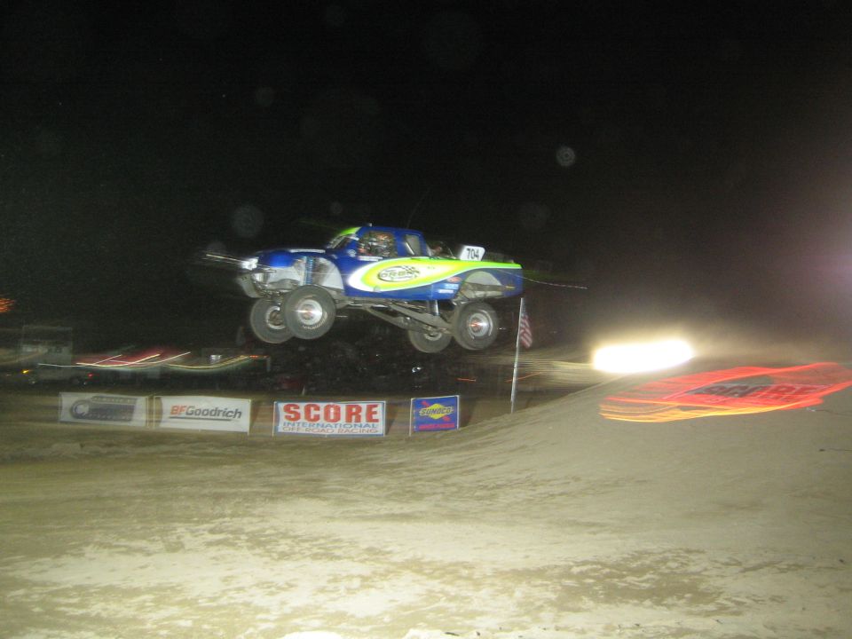 Blue and yellow off-road race truck jumping over a dirt mound at night past SCORE banners