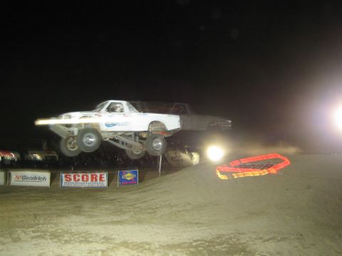 1990 Ford Bronco - white off-road race truck flies through the air off a dirt jump at night in front of SCORE and BFGoodrich banners.