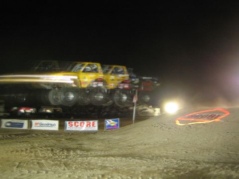 1990 Ford Bronco - yellow off-road truck blurs through a dirt jump at night, light streaking across SCORE and BFGoodrich banners.