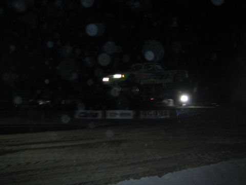 1990 Ford Bronco - green off-road truck jumps a dirt mound on a very dark night course, with bright headlights and lens spots in the frame.