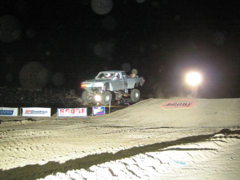 1990 Ford Bronco - gray off-road truck launches off a dirt jump at night in front of SCORE banners and an American flag.