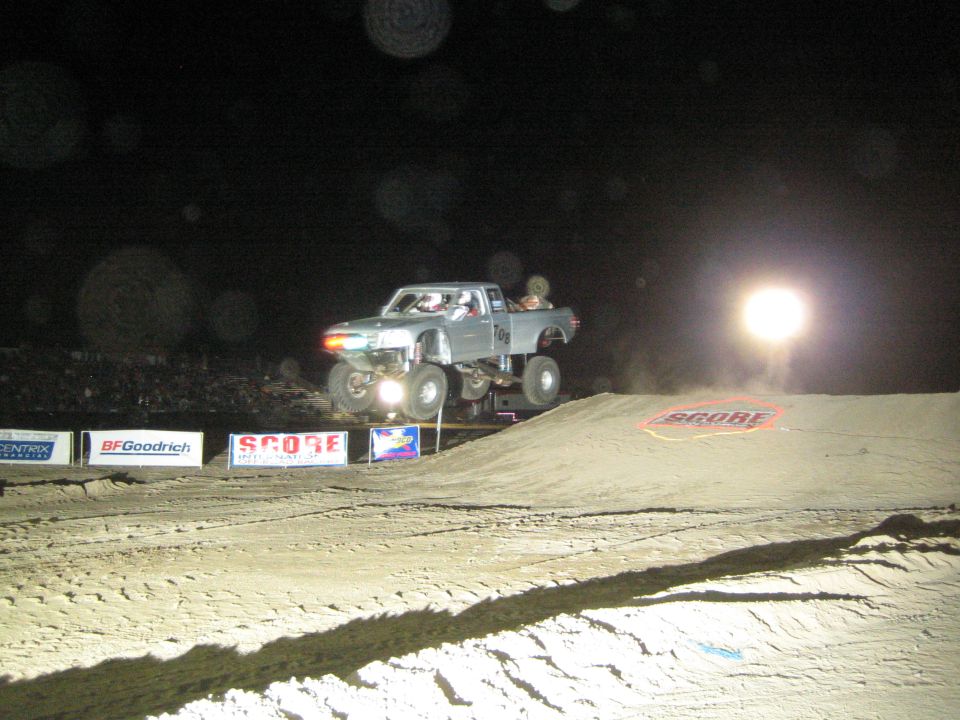Gray off-road truck airborne over a dirt jump at night past SCORE banners and a flag