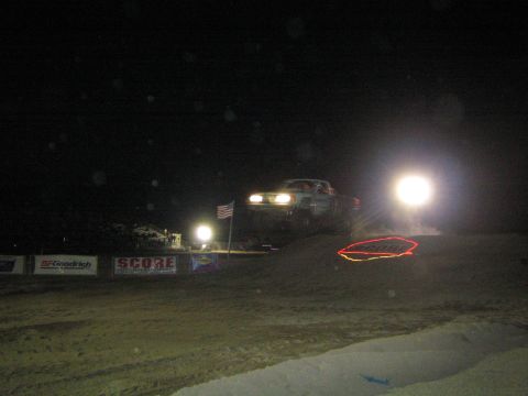 1990 Ford Bronco - off-road truck with headlights on crests a dirt jump at night near SCORE banners and an American flag, with a glaring light beside it.