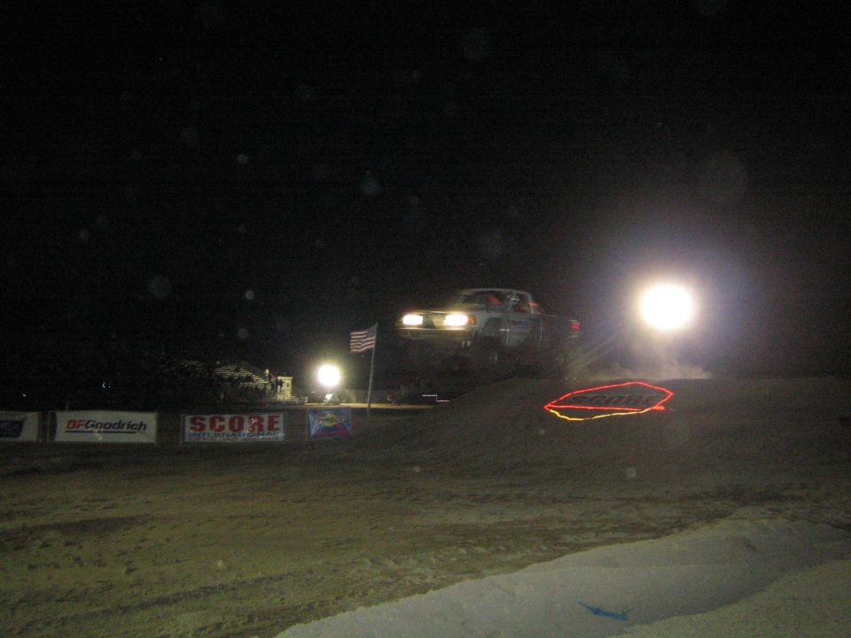 Off-road truck with headlights cresting a dirt jump at night past SCORE banners and flag
