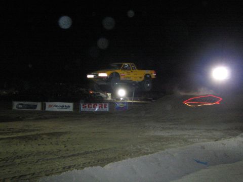 1990 Ford Bronco - yellow off-road race truck launches off a dirt jump under floodlights at a nighttime desert race with SCORE and BFGoodrich banners.