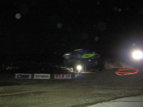 1990 Ford Bronco - green and black off-road racer hangs in the air over a dirt jump at night, lit by floodlights above sponsor banners.