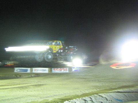1990 Ford Bronco - yellow off-road race truck flies off a dirt jump at night, headlights smeared into light streaks by motion, above SCORE banners.