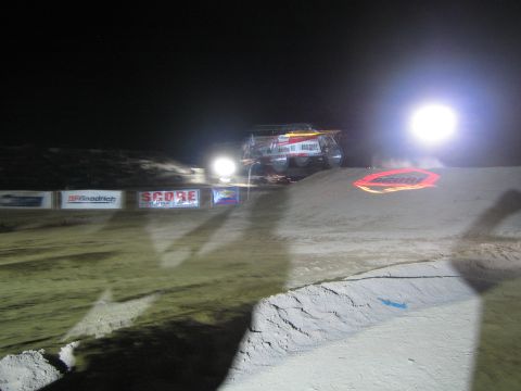 1990 Ford Bronco - off-road race vehicle crests a floodlit dirt jump at night behind a sand berm lined with SCORE sponsor banners.