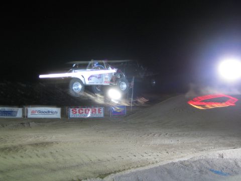 1990 Ford Bronco - white off-road race truck jumps a dirt mound at night, a bright headlight beam trailing across the floodlit course above SCORE banners.