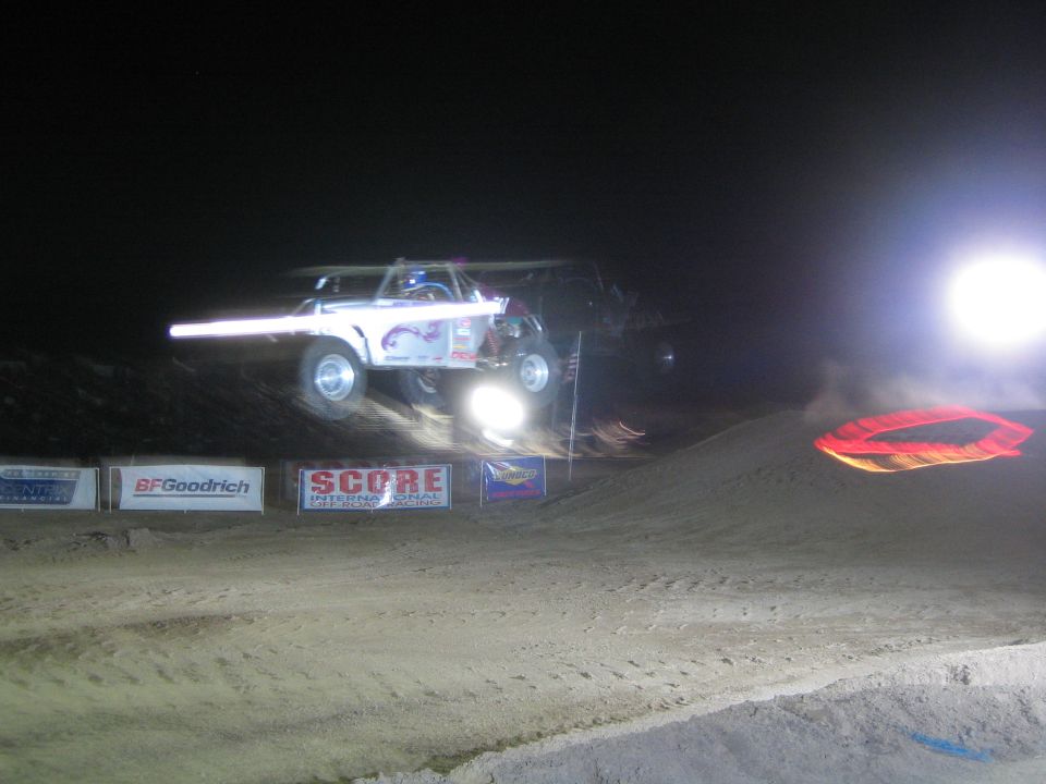 White off-road race truck airborne at night with headlight beam over dirt jump