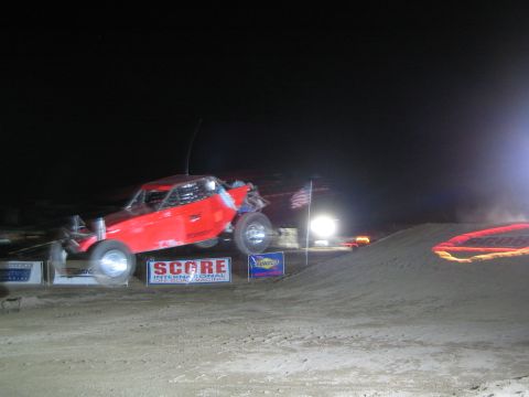 1990 Ford Bronco - red open-wheel off-road race buggy leaps off a dirt jump at night above SCORE banners under floodlights.